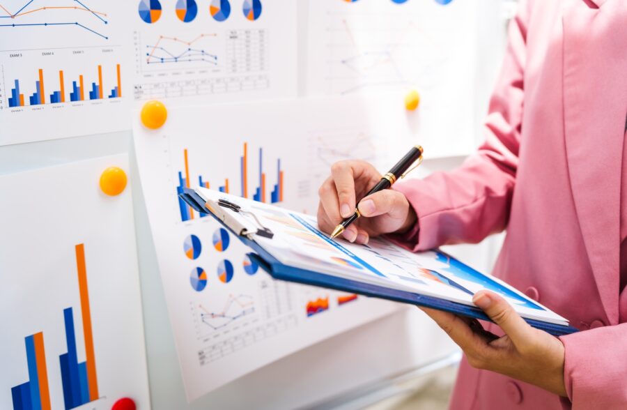 A businesswoman in a pink suit works at a desk with a whiteboard in the background.